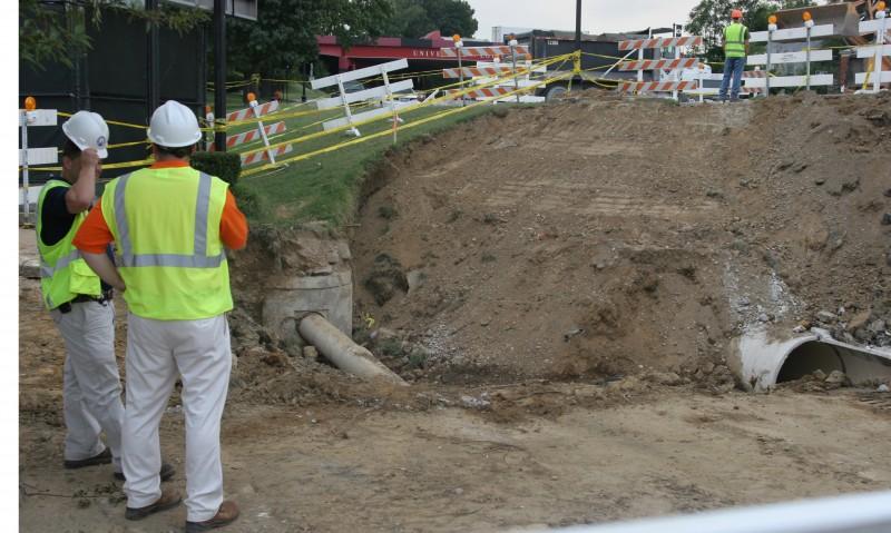 A worker in a hardhat and vest stands in front of the broken water main.